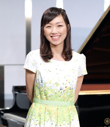 後ろ手を組み立って笑顔でこちらを向いているピアノデモンストレーター樋口紘子さんの写真