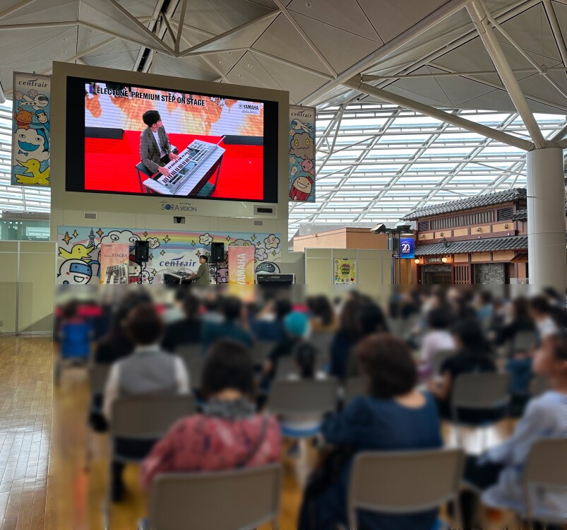 空港の大型ビジョンに演奏が映し出されたステージと観覧席の様子